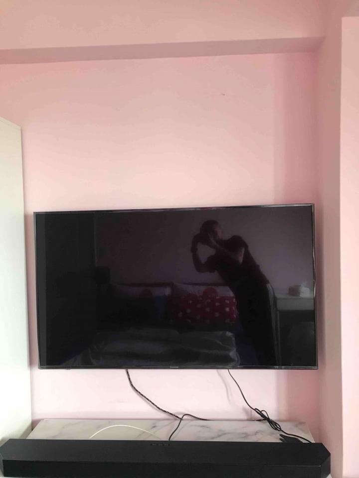 1) 照片顯示一個房間,牆壁是粉紅色的,牆上裝著一台黑色電視,下方有一台黑色的音響,電視上有著電線,電視螢幕呈現黑色,有一個人影反射在螢幕上