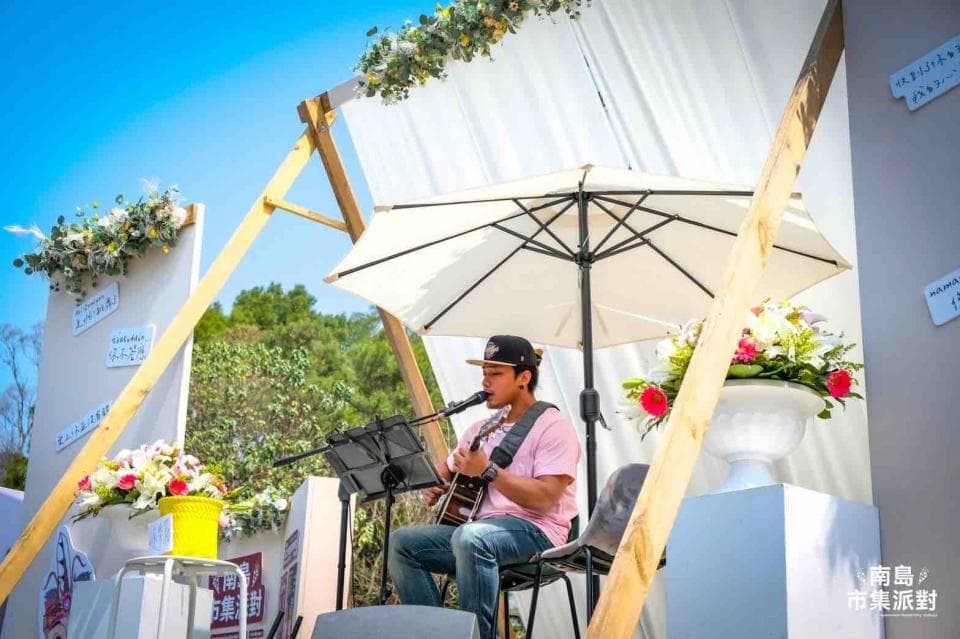 這張「戶外音樂表演」的照片是一個男人在舞臺上彈吉他唱歌,舞臺上放著鮮花和白色傘,背景是綠色的樹木和藍色的天空。