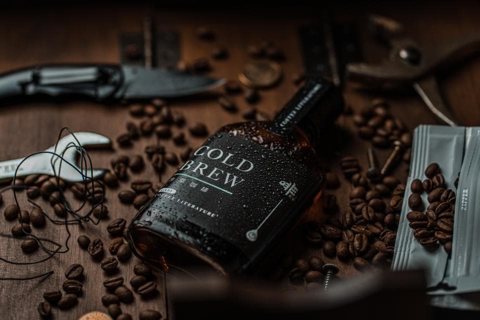 這張照片是一瓶標籤上寫著「COLD BREW」的咖啡酒,旁邊散落著咖啡豆。