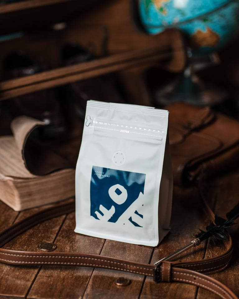 這張「咖啡豆包裝」的照片是一包白色包裝的咖啡豆,包裝上印著藍色的圖案,旁邊有一條棕色皮帶。