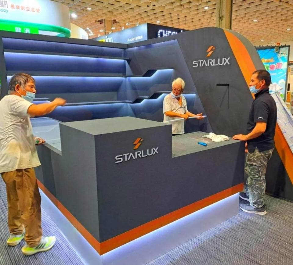 這張「星宇航空展臺」的照片是,展臺以灰色為主色調,上面印有星宇航空的標誌,並配有燈光效果,展臺上擺放了一些物品,有兩位工作人員正在工作,其中一位穿著白色工作服,另一位穿著深色工作服。