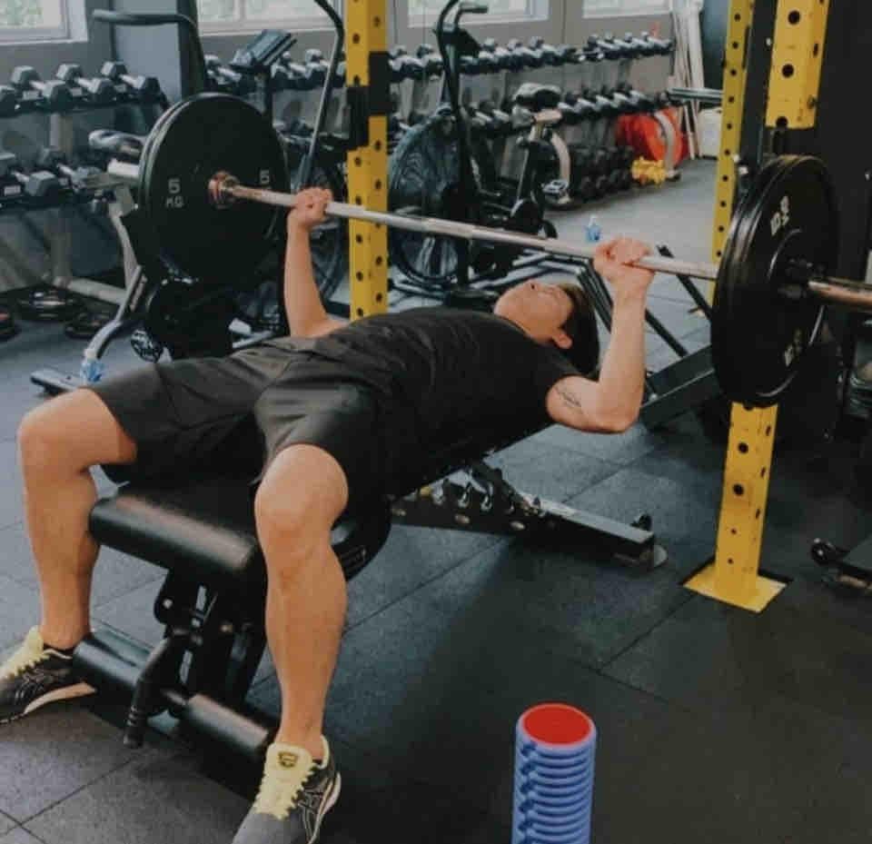 這張照片是一個男人正在健身房舉重。他正躺在一個臥推凳上,雙手握著槓鈴,準備舉起槓鈴。他表情專注,身體緊繃,顯然正在努力完成這項訓練。