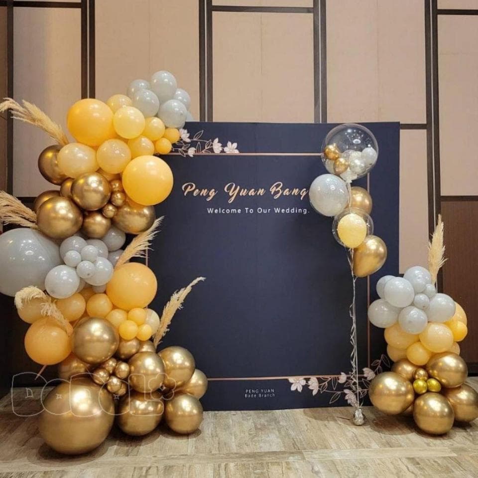 這張照片是一場婚禮的佈置,最重要的是用氣球做成的拱門。拱門由金色、銀色和黃色的氣球組成,中間還有一個透明的氣球,上面寫著「彭元邦,Welcome to Our Wedding」,非常浪漫。