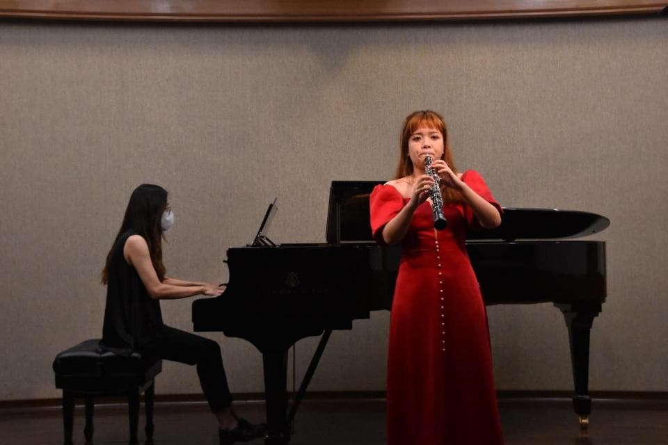 這張「音樂演奏」的照片是,一位穿著紅色長裙的女子正在吹奏雙簧管,一位戴著口罩的女子坐在鋼琴前準備彈奏。