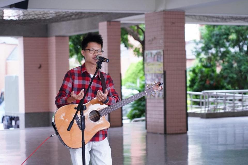 這張「年輕人彈吉他唱歌」的照片是一名男子在戶外唱歌,他穿著紅色的格子襯衫,戴著眼鏡,手拿吉他,並用麥克風唱歌。