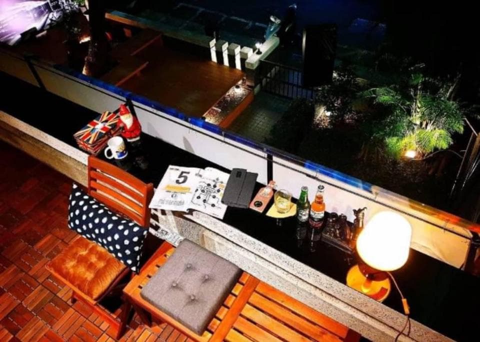 這張「戶外休息區」的照片是有一個木製的桌子,上面放著一些酒瓶、零食和一個燈,桌旁有兩個座位,背景是綠色植物和一個木質的平臺。