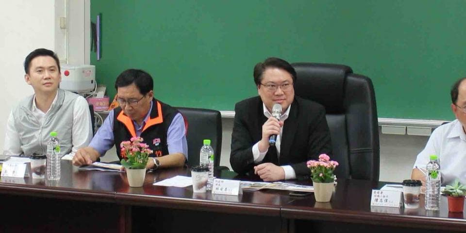 這張「會議」的照片是四個人坐在桌子旁開會,其中一個人拿著麥克風,桌上放著水杯、文件和盆栽。