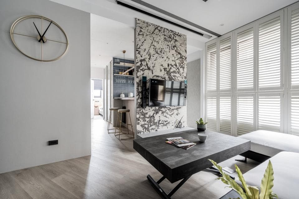 這張「現代簡約風格客廳」的照片是,其中有一個黑色大茶几、白色的沙發,以及一扇百葉窗。牆壁上有一面水泥牆,上頭裝飾著一個時鐘和電視。