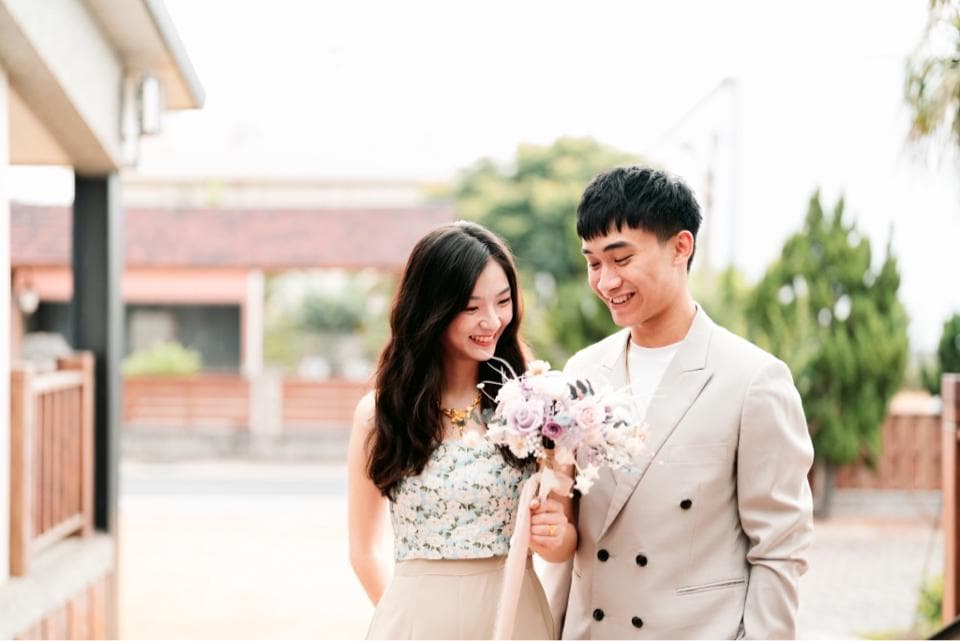 這張照片是一對情侶在戶外,男生穿著西裝,女生穿著白色洋裝,他們手拿著花束,看起來很甜蜜。