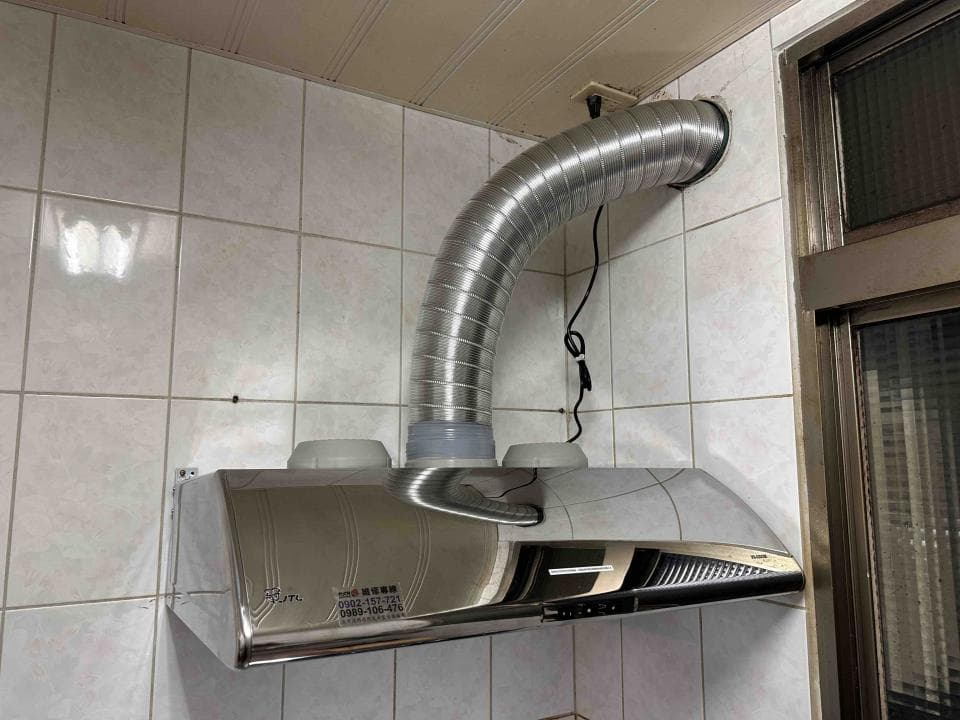 這張照片是一個廚房抽油煙機,最重要的部分是銀色的抽油煙機主體,它被安裝在牆上,並且連接了一個彎曲的排氣管。