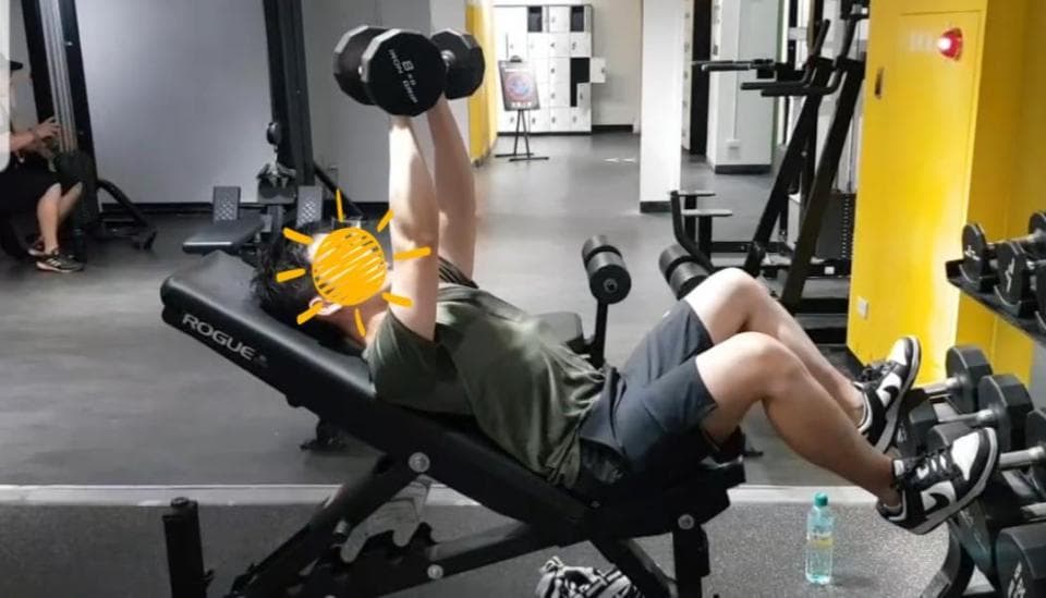 這張照片是一個男人在健身房舉啞鈴。他躺在一個黑色的健身椅上,雙手舉著啞鈴,雙腳踩在地上。他的臉上充滿著努力的表情,他的肌肉線條清晰可見。他周圍的健身器材也散發著一種強烈的運動氣息。