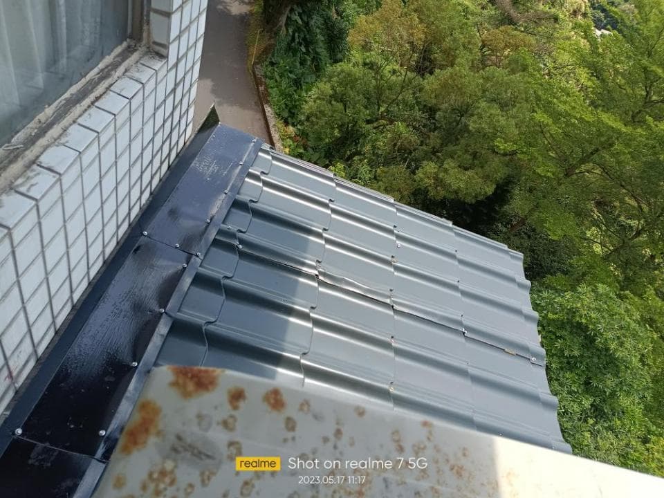 1) 照片顯示一個房屋的屋頂,屋頂是灰色的金屬瓦, 屋頂上有黑色的排水溝, 屋頂邊緣有白色磚塊牆, 房屋旁邊有綠色的樹木