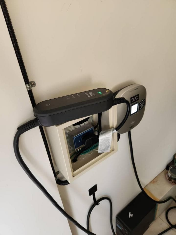 這張照片是一個電動汽車充電站,最重要的是充電槍,它是一個黑色的裝置,帶有電線和一個顯示屏。它連接到一個白色盒子里,盒子里有其他電氣元件。