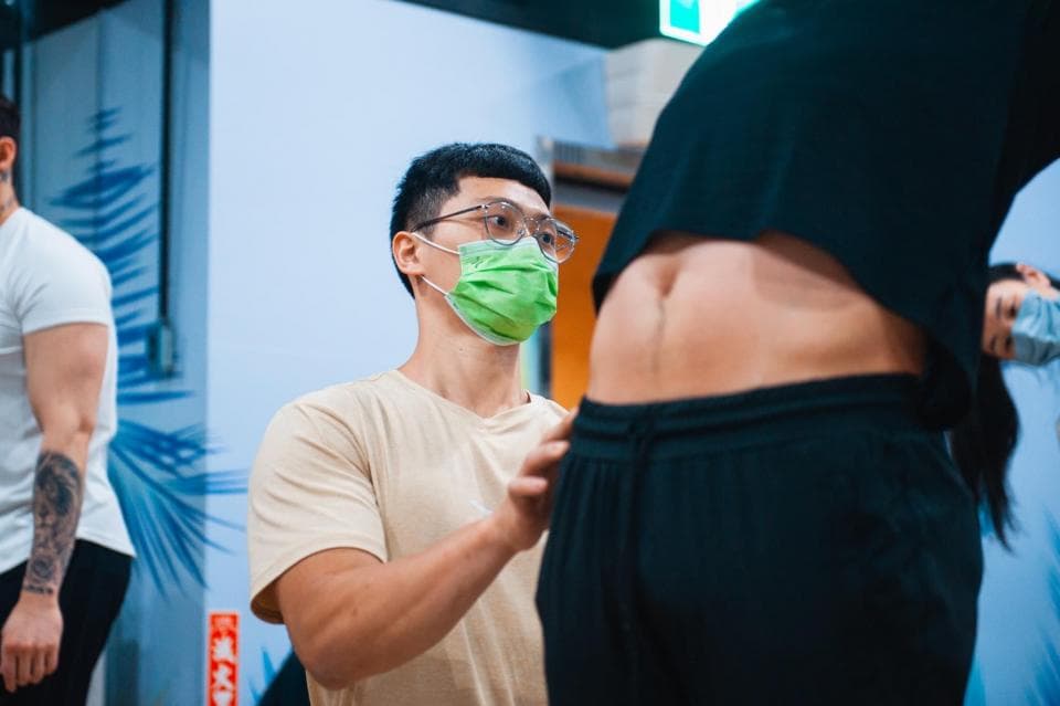 這張照片是一個戴著口罩的年輕人,他正在為另一個穿著黑色緊身褲的人調整衣服。