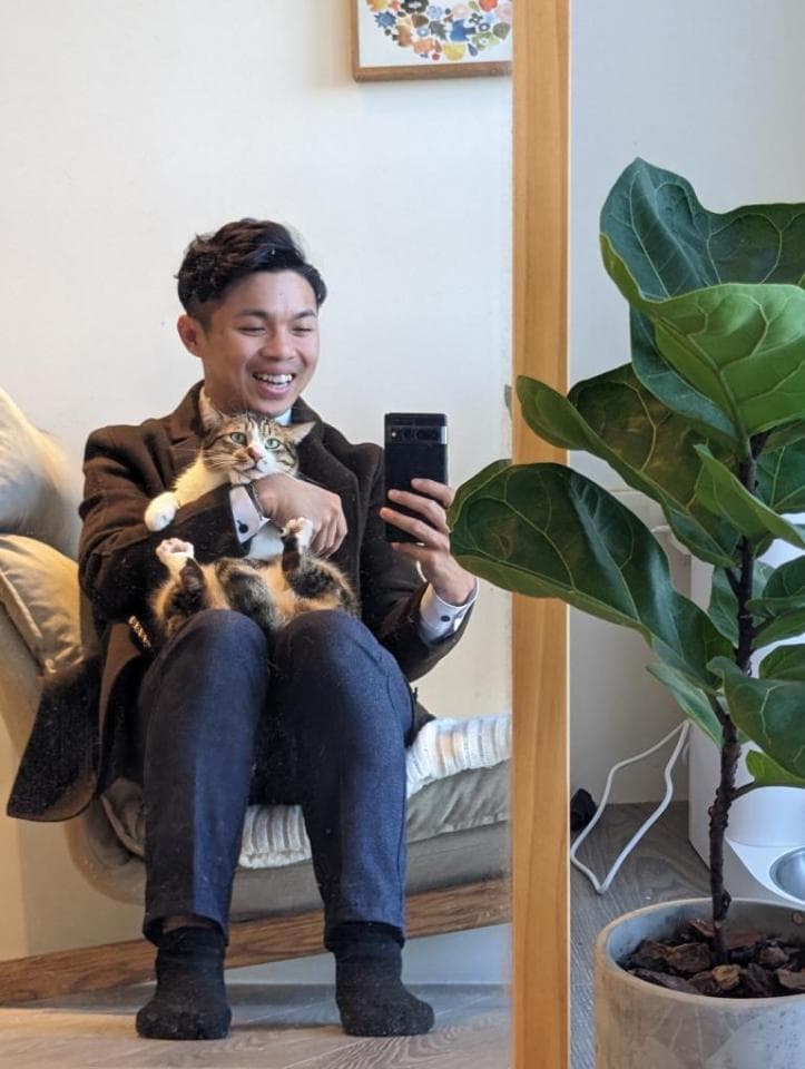 1) 照片顯示一位穿著西裝外套的男人坐在椅子上,他抱著一隻三色貓,並拿著手機自拍