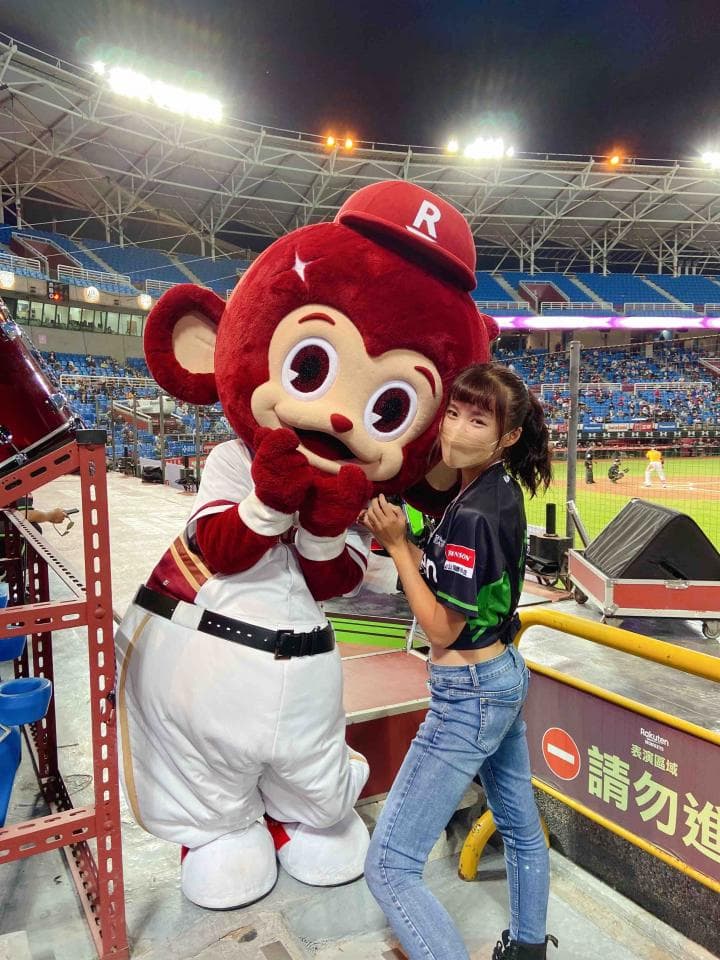 這張照片是一個穿著棒球制服的可愛吉祥物和一個年輕的女孩。吉祥物看起來像一隻猴子,戴著紅色的帽子,上面有字母「R」。女孩穿著黑色運動上衣和牛仔褲,她正和吉祥物拍照。