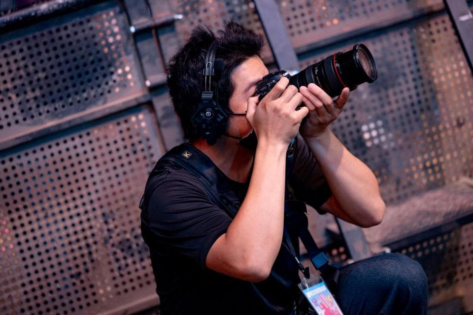這張「攝影師」的照片是一個男人在使用單眼相機拍照的畫面,他正用右手握住相機,並專注地看著鏡頭。他身穿黑色T恤,戴著耳機,表情認真。背景是一面鐵製的牆壁。