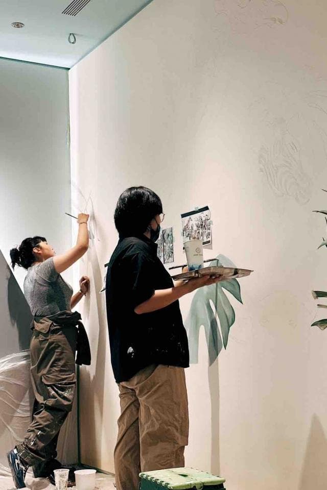 這張「兩個人在白牆上繪畫」的照片是,一個穿著黑色上衣的男生正在用刷子塗抹牆壁,另一個穿著灰色上衣的女生正在用刷子塗抹牆壁。