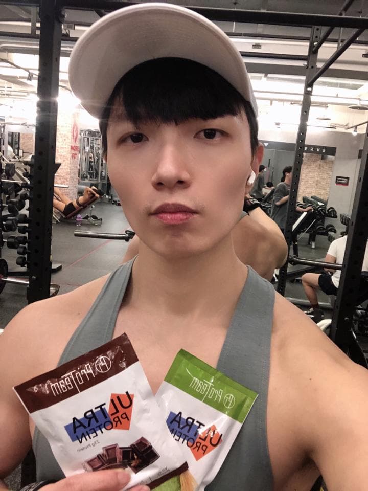 這張「健身房」的照片是一個戴著白色帽子、穿著灰色背心的年輕男子,他正在展示手中的兩包蛋白粉,他似乎在健身房裡運動完畢。