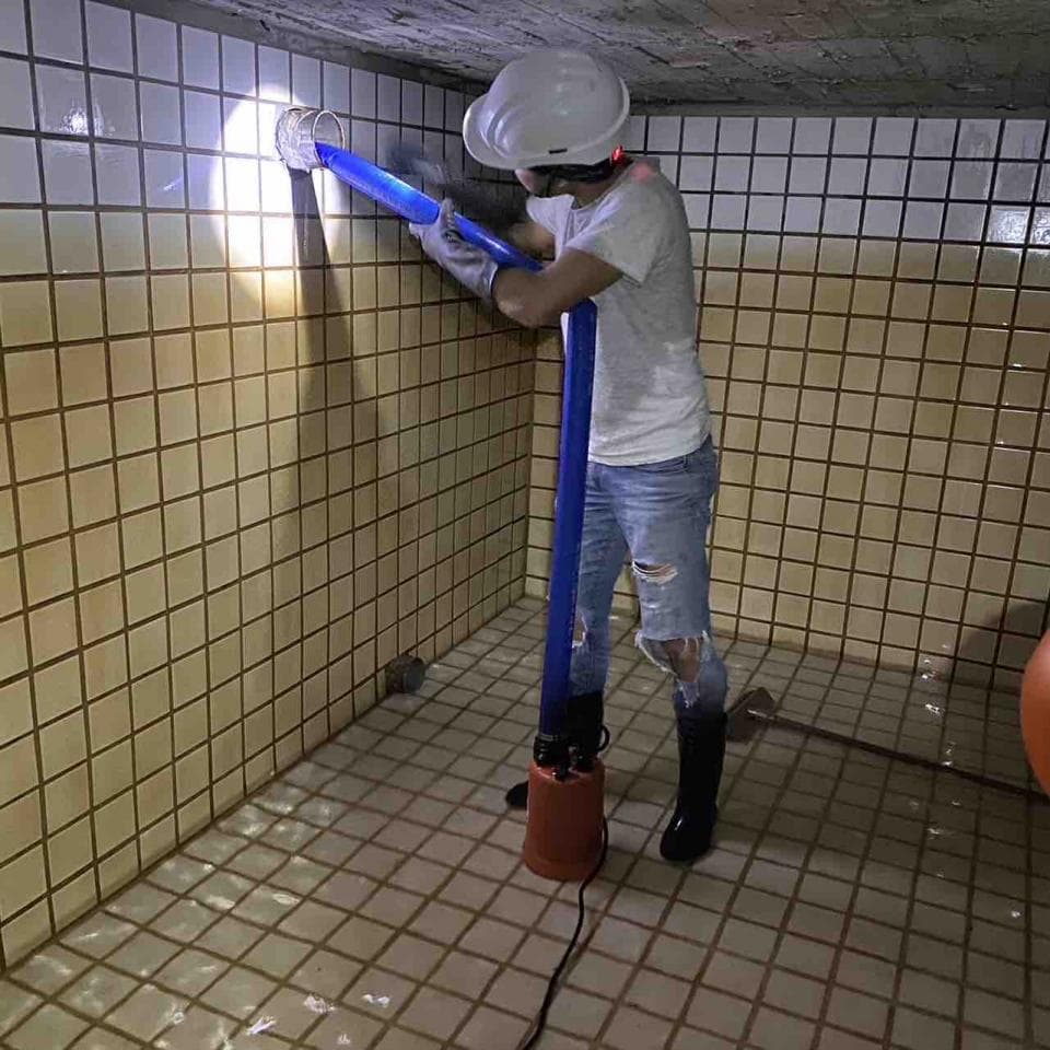 這張照片是一個工人正在使用抽水機排水,他站在被水淹沒的浴室裡。他穿著安全帽和雨靴,手持藍色抽水機,正在將水從浴室抽出去。