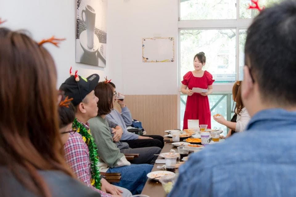 這張照片是一群朋友圍坐在桌子旁,其中一位女性正面向鏡頭,手持文件,似乎正在分享故事或演講。