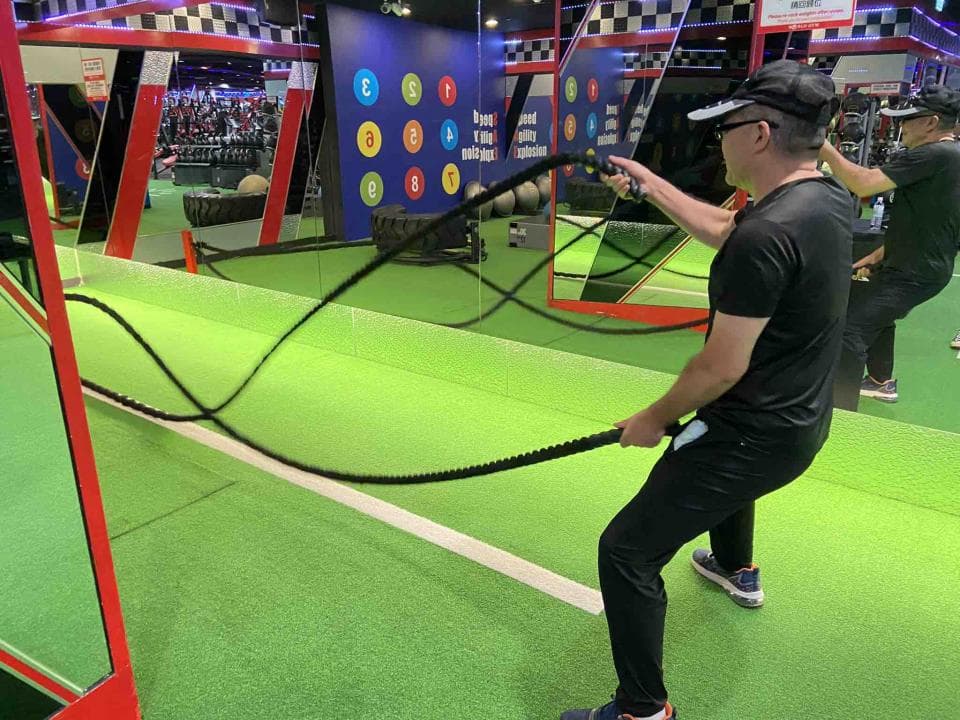 這張照片是一個健身房,照片中最重要的部分是一個男人正在用繩索做運動。他戴著帽子,穿著黑色運動服,雙手緊握著繩索,正專注地拉著繩索進行訓練。