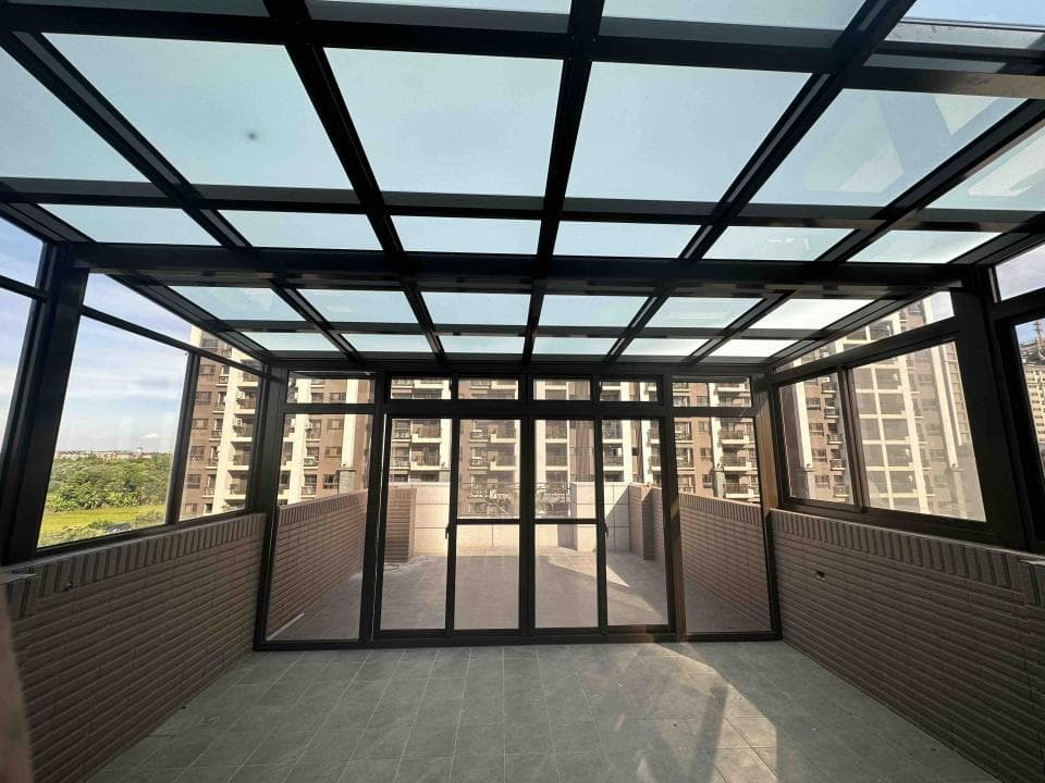 這張「陽臺加蓋」的照片是一個帶有玻璃屋頂的陽臺,陽臺有四面牆,其中一面是玻璃門,可以通往外面。陽臺上鋪著地磚,牆上裝飾著仿磚牆,看起來非常舒適。