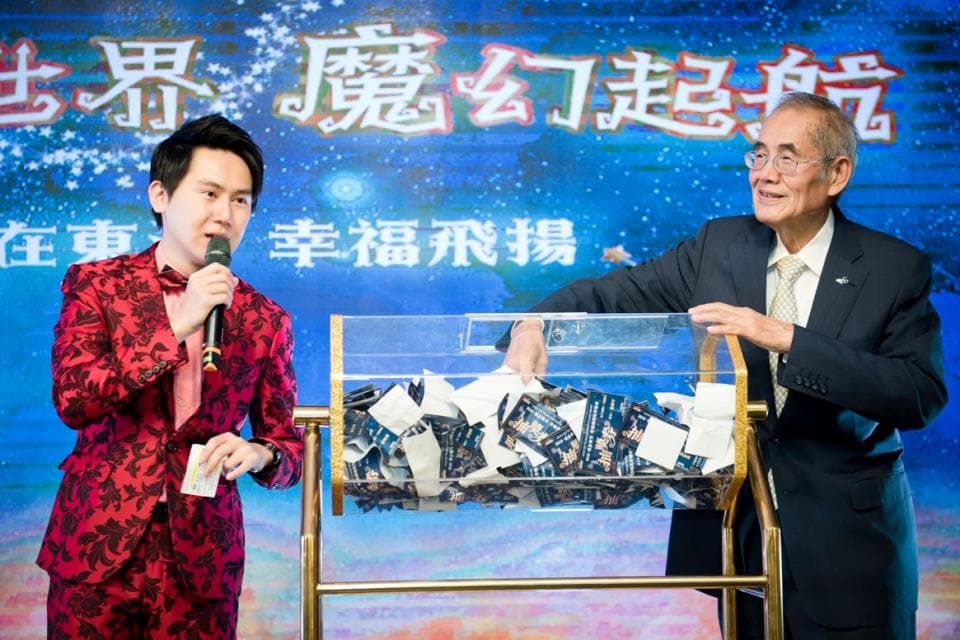 這張「抽獎活動」的照片是一個穿著紅色西裝的男子正在主持,一位穿著西裝的老人正在抽取獎品,他們站在一個透明的抽獎箱前。