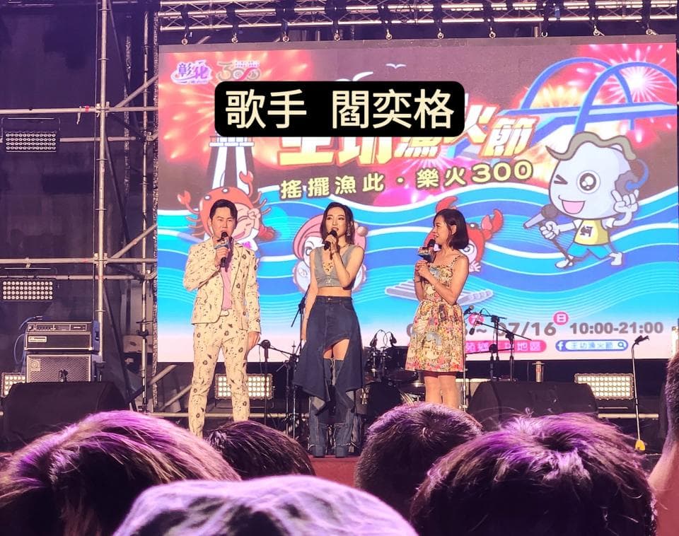 這張照片是一場音樂表演的現場,舞臺上有一位穿著花紋外套的男性歌手,兩位穿著不同風格服裝的女性歌手,他們正在臺上表演。