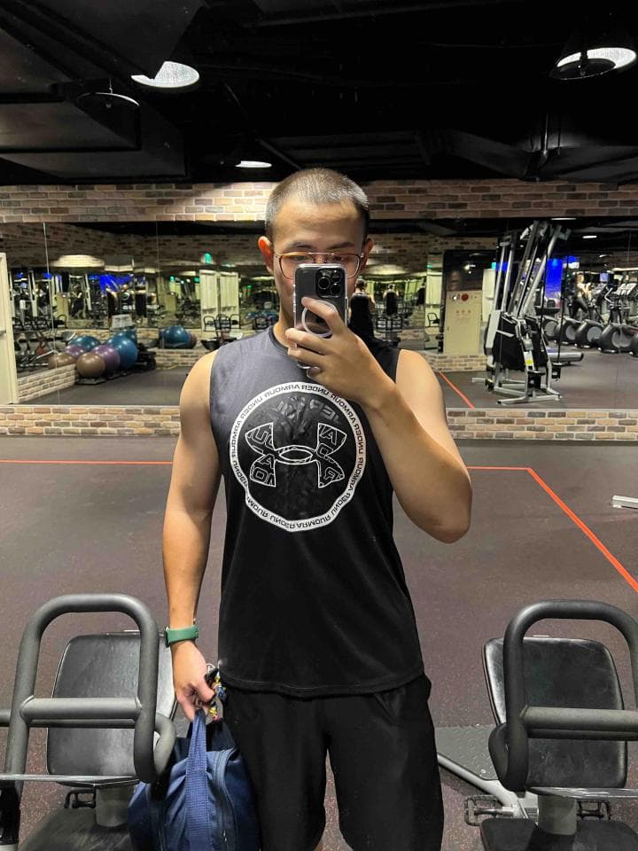 這張照片是一個穿著黑色運動背心,上面印著白色運動品牌logo的年輕男子,他站在健身房裡,拿著手機自拍。