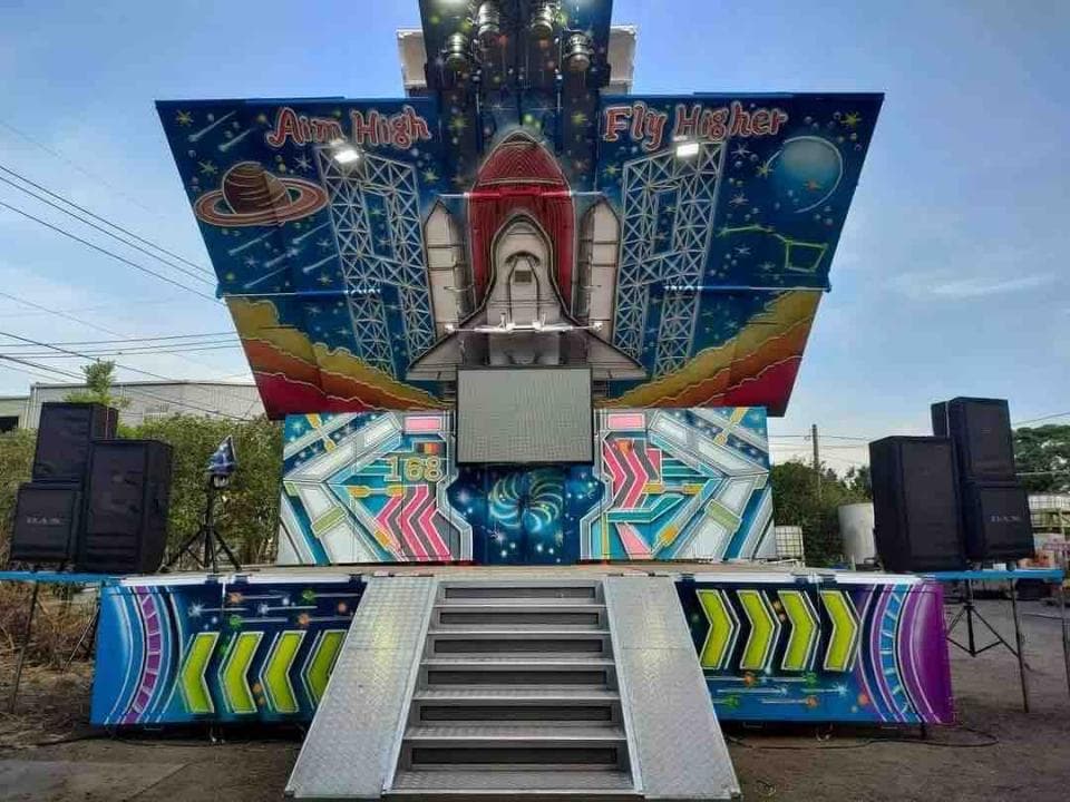 1) 照片顯示一座大型的戶外舞台,舞台上裝飾著太空梭和星星的圖案,並寫著 "Aim High" 和 "Fly Higher" 的標語