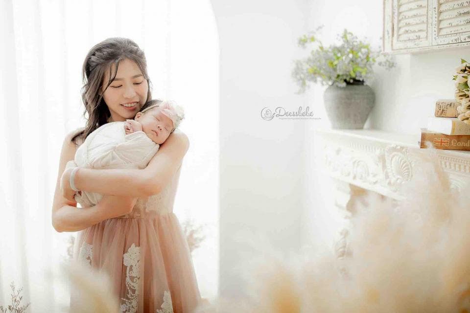 1) 照片中是一位年輕女性穿著粉紅色蕾絲洋裝,懷抱著一名嬰兒,背景是白色牆壁,角落擺放著花瓶和裝飾品
