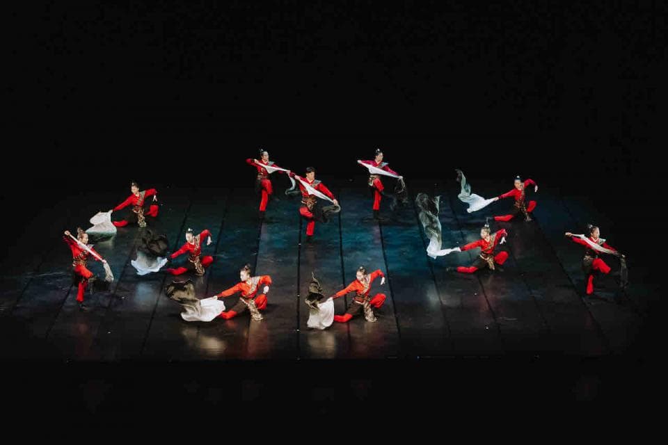 這張照片是一群身穿紅色服裝的舞者在舞台上表演傳統中國舞蹈,他們手持白色絲綢,在黑色背景下優雅地跳躍,展現了中國文化的傳統美感。
