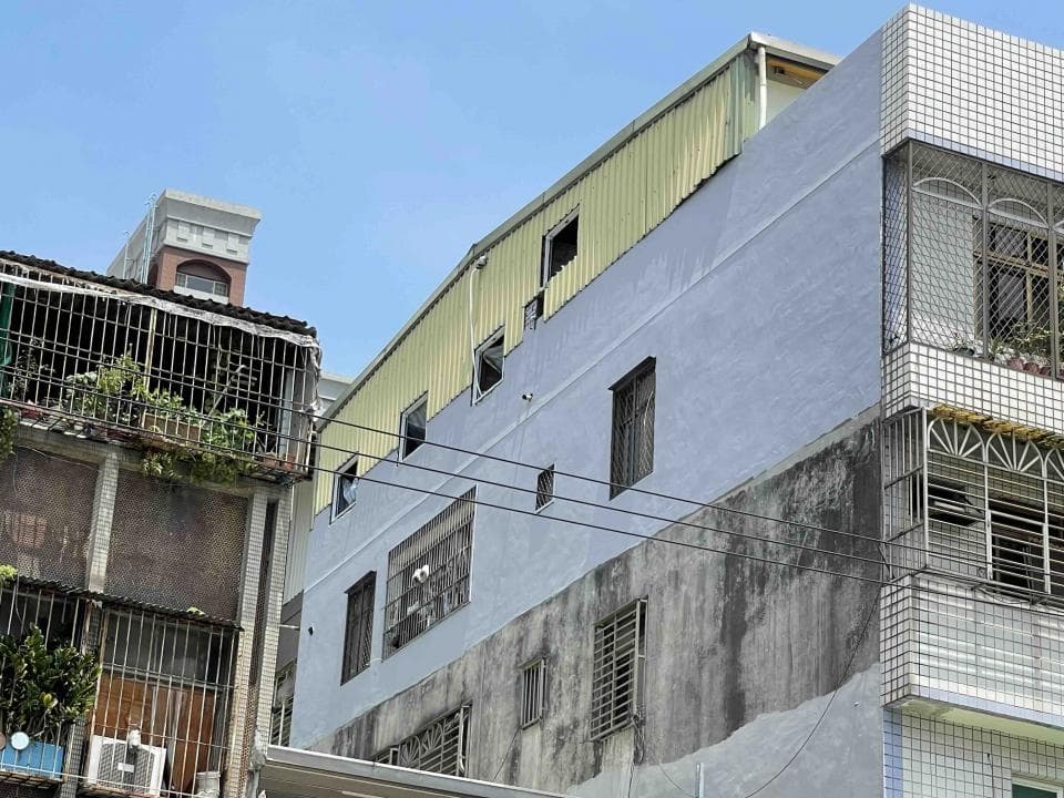 1) 照片顯示兩棟建築物,較大的建築物左側有金屬屋頂,右側有水泥牆,牆上有些污漬,建築物正面有許多窗户,窗户有鐵窗