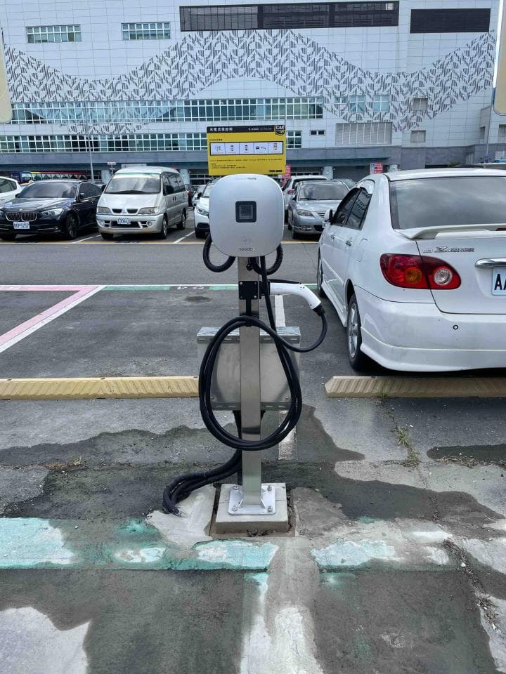 1) 照片顯示一個電動汽車充電站,充電站位於停車場旁,後面停著多輛汽車,建築物前方有一塊標誌牌
