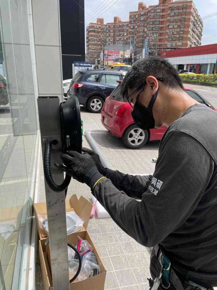 這張照片是一個男人正在安裝牆壁上的黑色裝置。他穿著黑色襯衫,戴著黑色手套,專注地操作著。