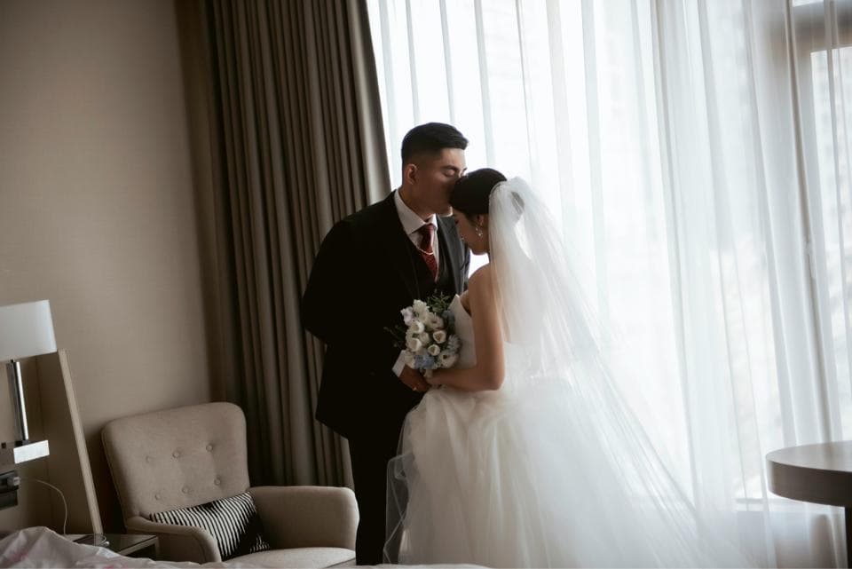 這張「婚禮」的照片是一對新人在房間裡,新郎親吻新娘的額頭,新娘穿著白色婚紗,新郎穿著黑色西裝,他們手裡拿著花束,背景是窗戶和窗簾。