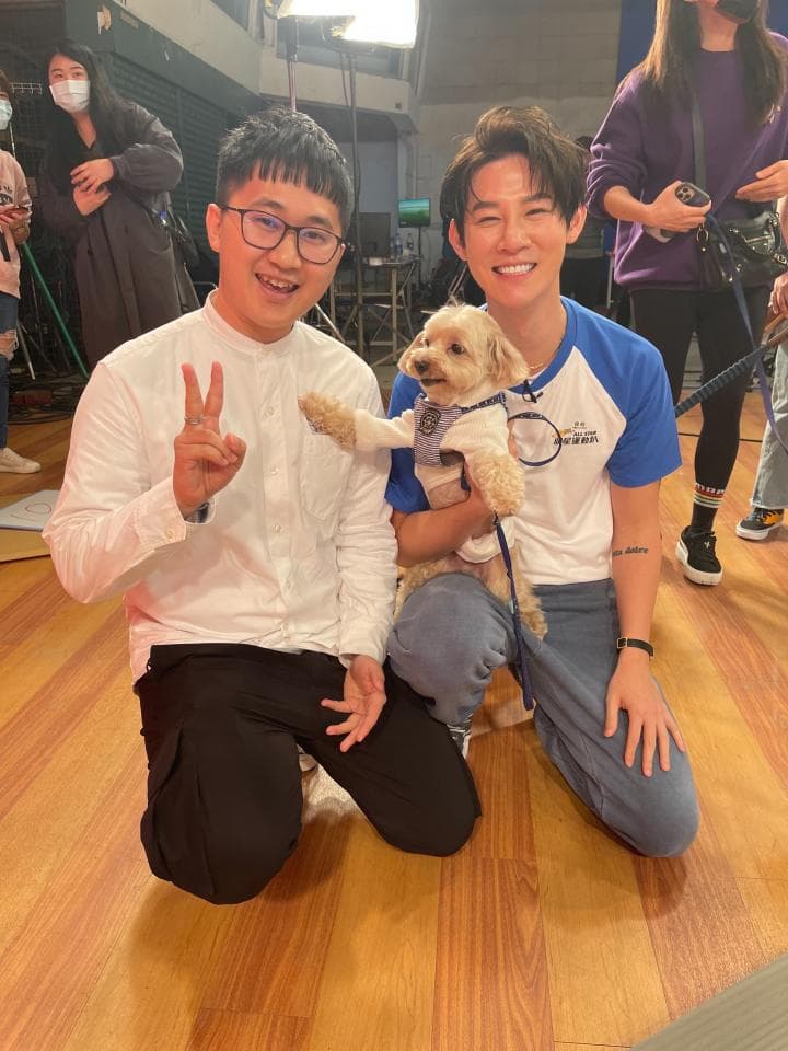 這張照片是一隻可愛的小狗被兩位男生抱著。狗狗毛色金黃,看起來很溫順可愛,兩位男生都露出燦爛的笑容,看起來很開心。