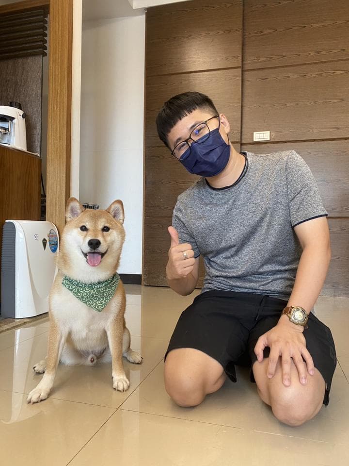 這張照片是一個戴著口罩的男人,他正跪在地上,用右手比著讚,他的左邊有一隻戴著綠色圍巾的柴犬。