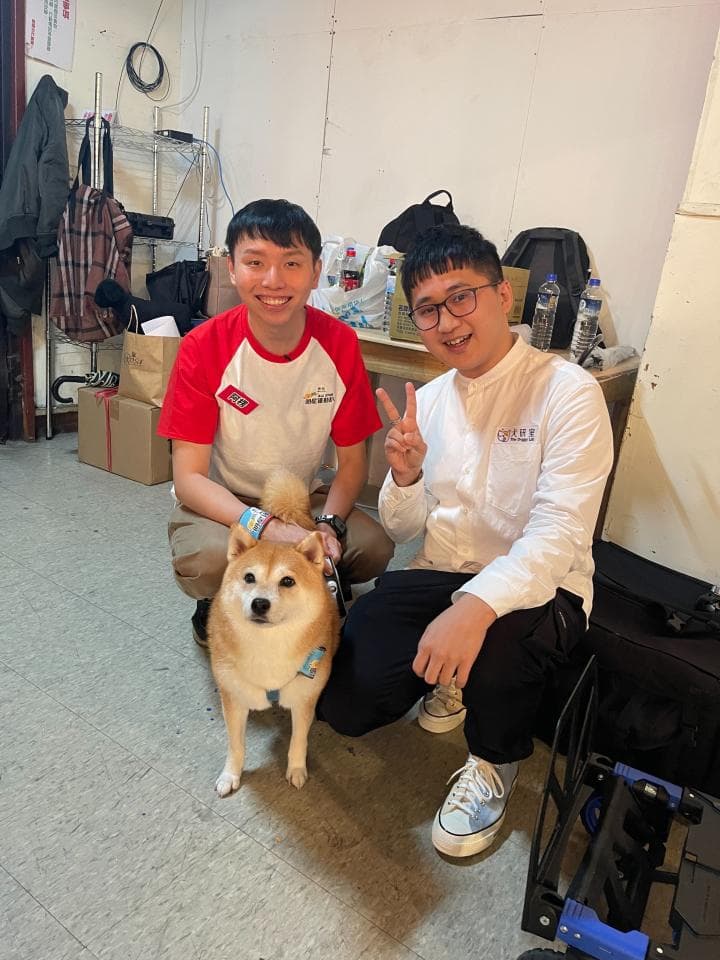 這張照片是兩個男人和一隻柴犬的合照,他們坐在房間裡,柴犬坐在地上,其中一個男人正用手輕輕地撫摸著柴犬的頭。