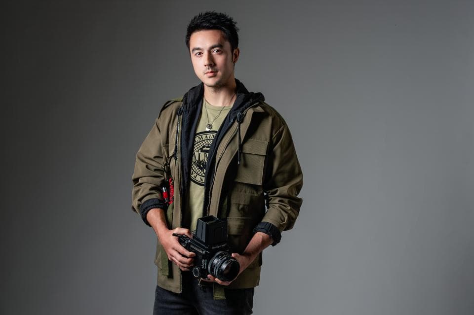 這張「男子穿著綠色外套,手持相機」的照片是,一個年輕男子站在灰色背景前,他穿著一件綠色夾克,裡面穿著一件綠色T恤,他手裡拿著一臺相機,看起來很自信。