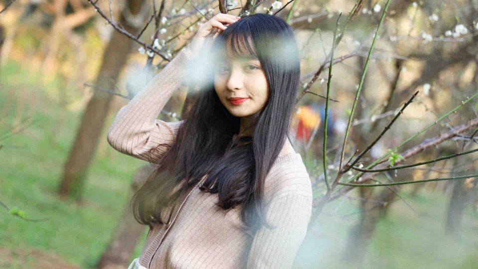 這張照片是一個年輕女子站在樹叢中,她穿著米色的針織衫,長長的黑色頭髮,用手輕輕地撫摸著她的臉。她看起來很安靜,眼神充滿著沉思。背景是綠色的草地和樹枝,讓她看起來更加美麗。