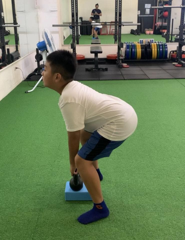 這張照片是一個年輕人在健身房舉重。他正準備舉起一個黑色的壺鈴,他的姿勢非常正確,腳踩在一個藍色的墊子上,顯然他正在努力鍛鍊自己的力量和肌肉。