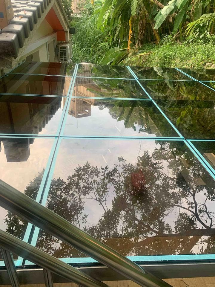 這張照片是一個玻璃屋頂,屋頂的玻璃呈現出藍色的色調,反射出周圍環境的景象,包括樹木和建築物。玻璃屋頂是由多個玻璃面板組成,並以金屬框架固定。屋頂的下方是金屬欄杆,欄杆呈銀色,與玻璃屋頂的顏色形成對比。