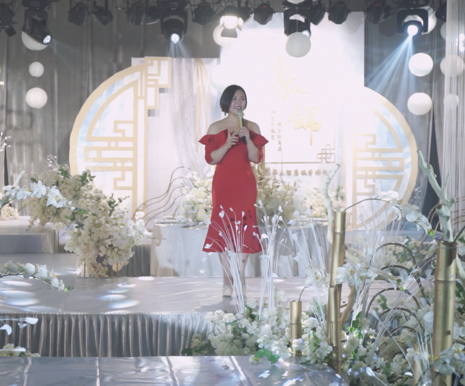 這張「婚禮」的照片是一個穿著紅色禮服的女人站在舞臺上,背景是白色的花朵和金色的裝飾,旁邊還有金色柱子。