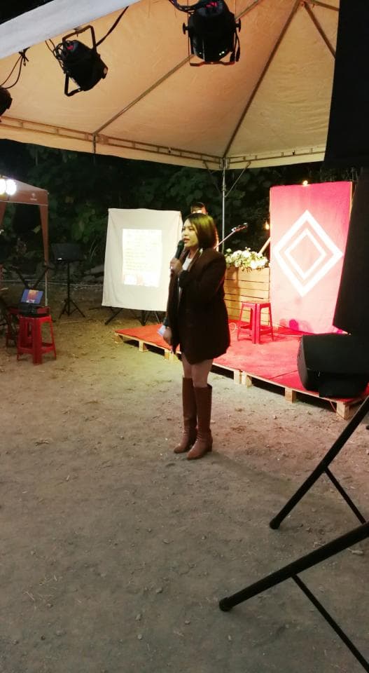 這張「戶外活動」的照片是一個女人正在臺上說話,她戴著耳機,穿著棕色外套和高筒靴,背景是一塊紅色布幕和一個白色布幕。