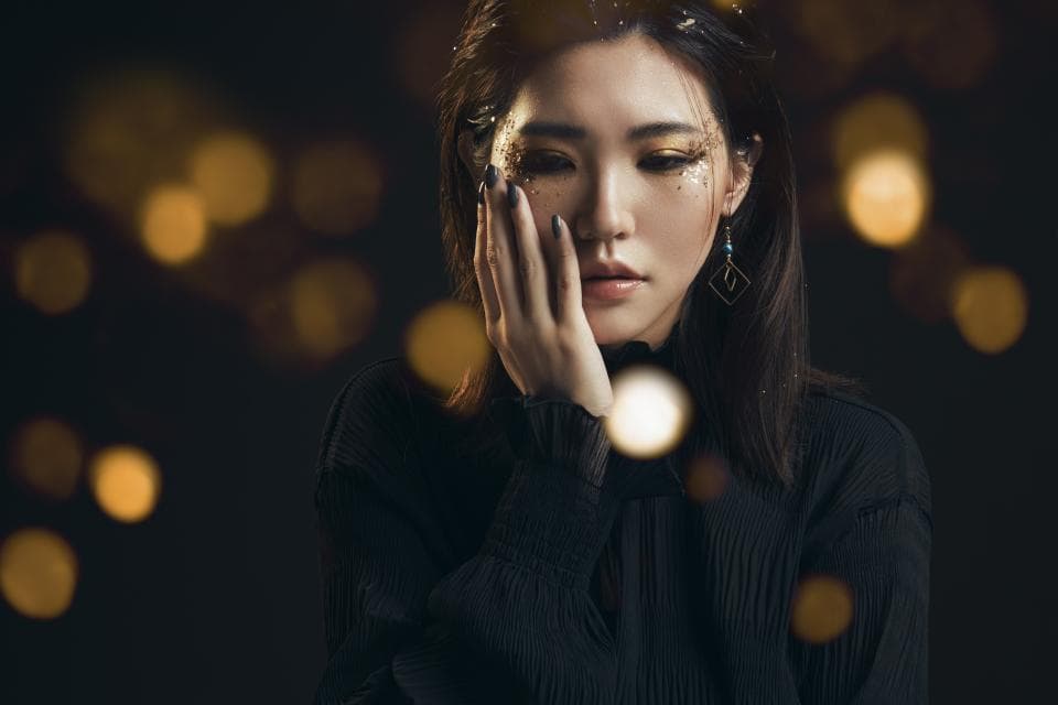 這張照片是一個穿著黑色毛衣的女生,她低頭看著她的手,似乎在沉思或思考著什麼。她戴著金色的耳環,她的臉上也有金色的亮片,這讓她看起來更加神秘和迷人。