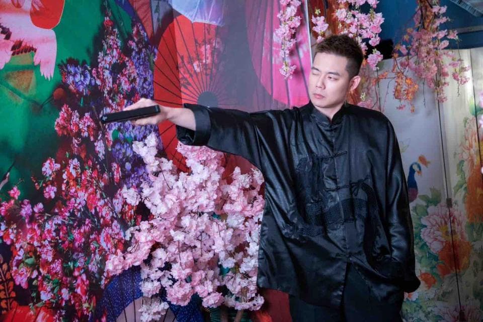 這張照片是一個穿著黑色唐裝的年輕男子,他正在手持一個黑色物體,背景是粉紅色的花朵和紅色、綠色的裝飾。