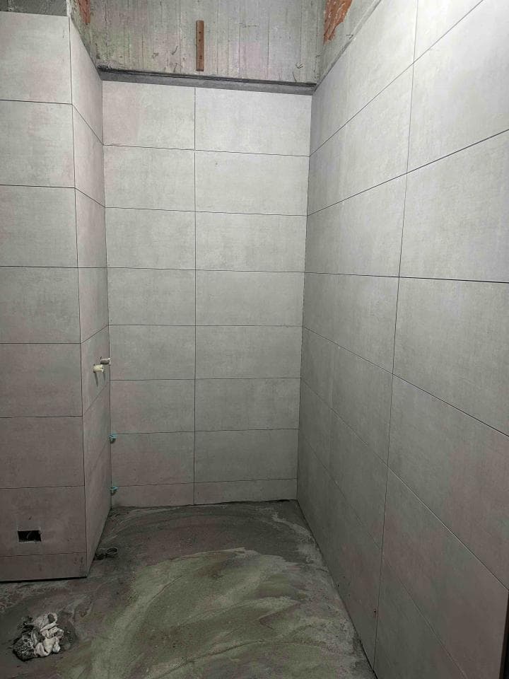 1) 這張照片顯示一個正在施工中的浴室,牆壁上鋪著淺灰色的瓷磚,地面上鋪著水泥砂漿,尚未完成