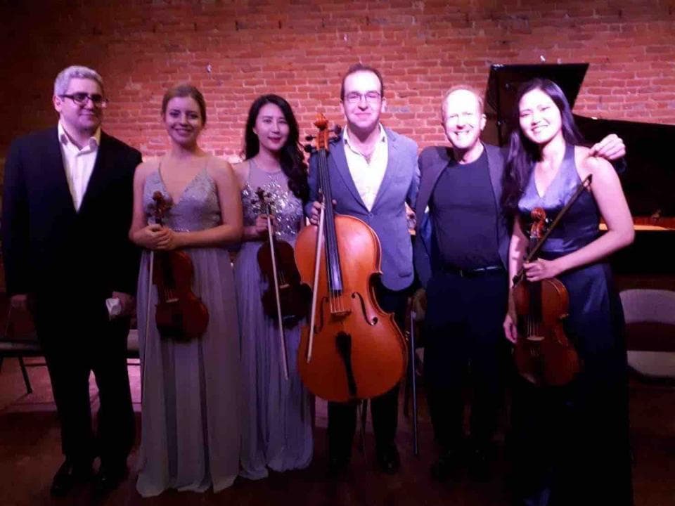 這張照片是一個室內音樂會,照片中的五位音樂家正在演奏小提琴和中提琴。其中一位音樂家正在演奏大提琴,他的位置在照片的中心,他穿著深色西裝,看起來非常專注。